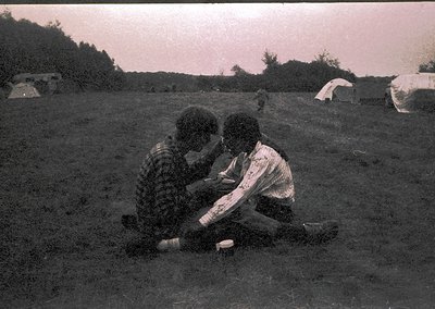 Vintage black-and-white photo of two individuals sitting closely in a grassy field, possibly mid-20th century. One wears a pa...