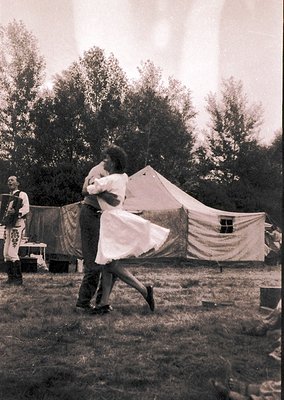 Couple dancing outdoors in vintage tented campground, mid-20th century. Man in dark trousers leads woman in flowing white dre...
