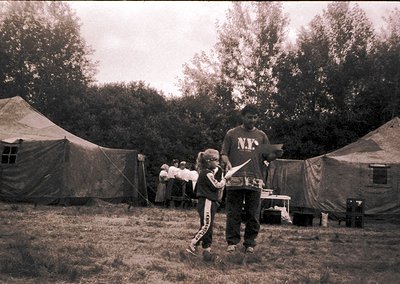 Vintage black-and-white photo of a makeshift campsite with canvas tents in a wooded area. A man in a hoodie and child in a tr...