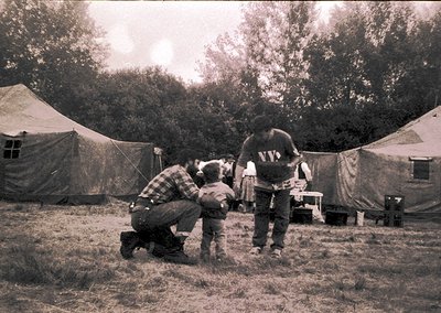 Black-and-white photo of a temporary encampment with beige canvas tents in a grassy, wooded area. Three adults and a child st...