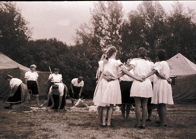 Mid-20th century outdoor gathering with men in rolled-sleeve shirts and women in knee-length dresses. Group stands near a mak...