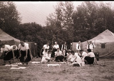 Vintage group engaged in traditional rope-making outdoors, seated on grass under large tents. Clothing suggests early-to-mid ...