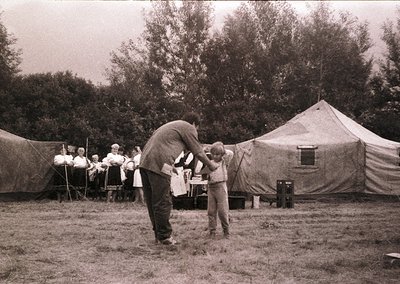 Military-style canvas tents in a grassy outdoor setting, mid-20th century. A man in a dark uniform adjusts a child’s cap, whi...
