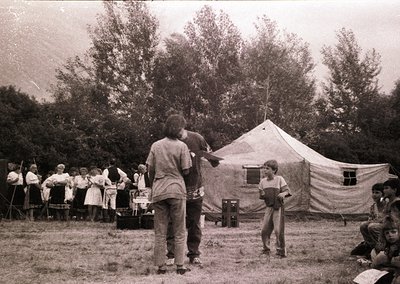 Vintage black-and-white photo of a group camping event in a grassy clearing. Central tent features a red cross emblem, sugges...