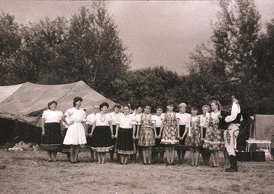 Vintage group photo of women in traditional folk attire, likely Eastern European, holding hands in a circle. A man in a disti...