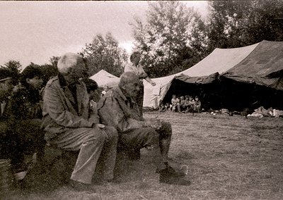 Two elderly men converse outdoors under makeshift tents in a rural setting, likely mid-20th century. Clothing suggests tradit...
