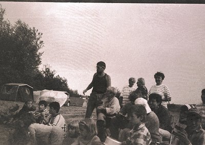 Group of people gathered outdoors in a grassy area, likely a 1970s camping scene. Central figure in sleeveless shirt poses wi...