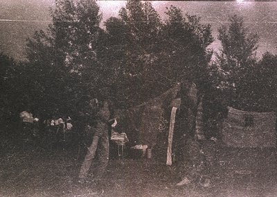 Vintage sepia-toned photo of a rural campsite with a lone figure seated near a small wooden table and chair. Surrounding foli...