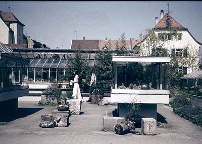Vintage garden pavilion with mid-century modern design, featuring a glass-enclosed greenhouse and stone planters. Two women i...
