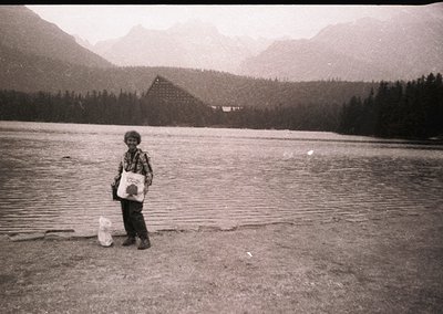 Child stands by a serene alpine lake, holding a white plastic bag and a cylindrical container, likely for fishing or collecti...