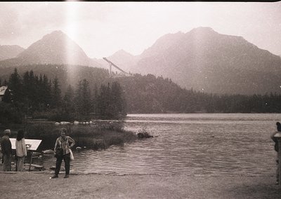 Black-and-white shot of a mountainous lakeside scene with a suspension bridge in mid-construction, surrounded by dense forest...