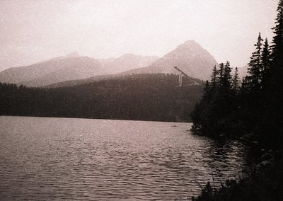 Black-and-white alpine lake framed by dense coniferous forest and jagged peaks, evoking early 20th-century travel photography...