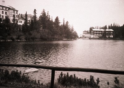 Vintage black-and-white lakeside resort with two-story wooden buildings surrounded by dense pine forest. Reflective water and...