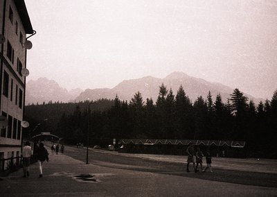 Mid-century alpine resort scene with multi-story concrete building and forested mountain backdrop. Snow-capped peaks frame th...
