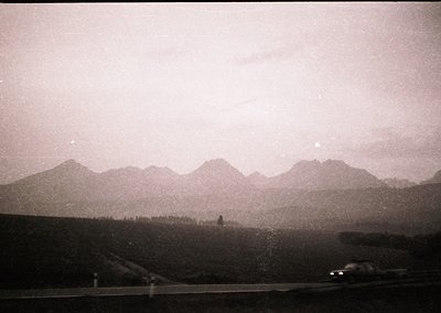 Vintage black-and-white shot of a lone car driving along a winding alpine road, flanked by dense conifer forests and jagged p...