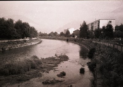 Mid-20th century urban river scene with concrete embankments and sparse vegetation. Low-rise residential buildings and indust...