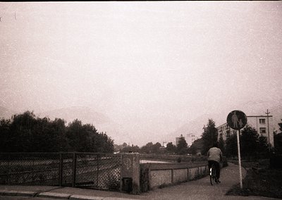 Black-and-white street scene featuring a lone cyclist crossing a rusted metal bridge over a body of water. Surrounding greene...