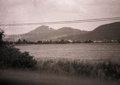 Vintage sepia-toned landscape featuring rolling hills, sparse vegetation, and a distant village. Power lines frame the top ed...