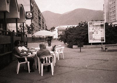Three men seated at an outdoor café table under a white umbrella in a mid-20th-century urban setting. Concrete apartment bloc...