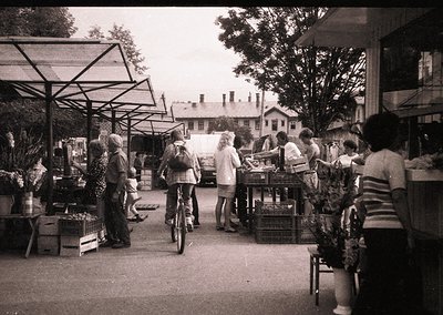 Vintage black-and-white street market scene with wooden stalls displaying fresh produce, baskets, and crates. People in 1970s...