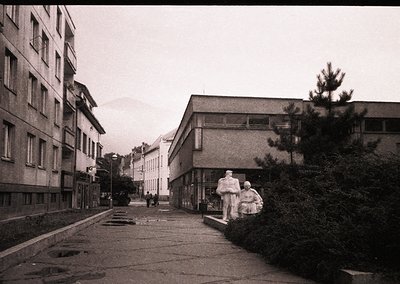Mid-century urban street with Soviet-era Brutalist architecture—concrete apartment blocks and low-rise institutional building...
