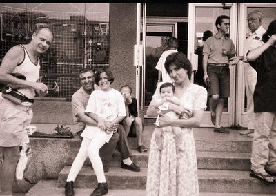 Black-and-white candid shot of a lively 1970s gathering outside a building with industrial scaffolding. Central figures: a wo...