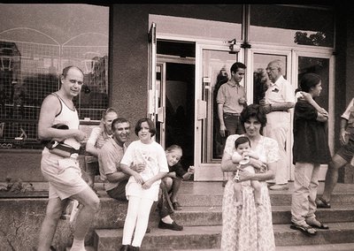 A candid 1970s-era black-and-white photo captures a lively gathering outside a Soviet-era apartment building. A diverse group...