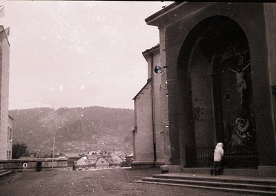 Black-and-white street scene featuring a weathered religious archway with a crucifix and saint mural. A lone figure in a long...