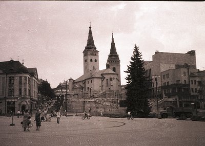 Mid-20th century street scene featuring a prominent church with twin spires and a central tower, likely European. Pedestrians...