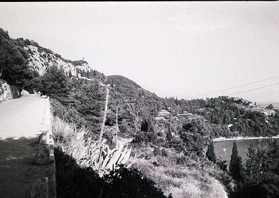Black-and-white coastal roadside scene with rugged cliffs and dense vegetation. A lone utility pole and power lines stretch a...