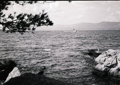 Black-and-white coastal scene featuring turbulent waves crashing against jagged rocks. Distant sailboat glides on calm waters...