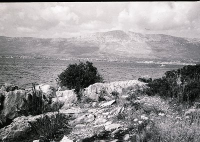 Black-and-white coastal landscape featuring rugged rocky foreground with sparse vegetation. Distant shoreline curves around a...