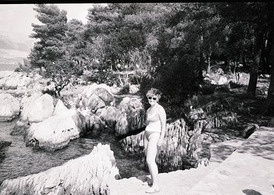 Black-and-white shot of a woman posing by a natural rock waterfall in a lush, forested setting. Mid-20th century attire (biki...