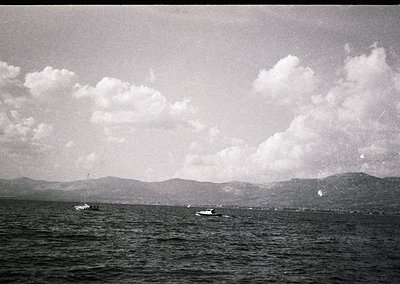 Vintage black-and-white seascape featuring two small boats on calm waters, with distant mountainous coastline under scattered...