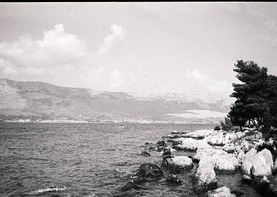 Mid-20th century black-and-white coastal scene: jagged rocks frame a rocky shoreline with minimal snow, while a lone boat dri...