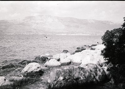 Vintage black-and-white coastal scene featuring jagged rocks in foreground, a lone boat on choppy waters, and misty mountains...