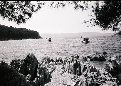 Black-and-white coastal scene featuring jagged rock formations in the foreground and calm waters with three small boats ancho...