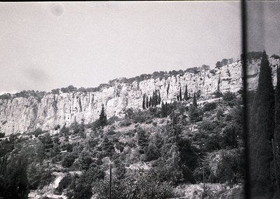 Mid-20th century black-and-white shot of rugged limestone cliffs with sparse vegetation. Narrow winding road hugs base of ste...