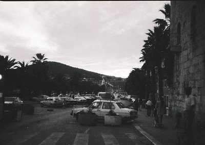 Mid-century coastal street scene with vintage cars parked along a promenade. Palm trees line the road, and a stone building o...