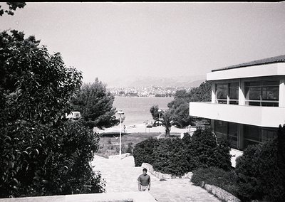 Mid-century seaside resort building with flat roof and large windows overlooking a calm coastal inlet. A man sits on stone st...