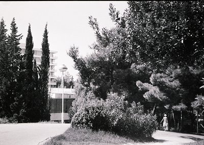 Mid-century modern seaside resort building with cylindrical balconies, framed by dense Mediterranean foliage. A lone figure i...