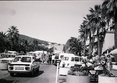 Mid-20th century seaside promenade with vintage cars, palm trees, and Mediterranean architecture. Crowded pedestrian area wit...