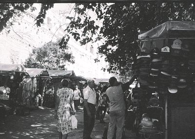 Vintage black-and-white outdoor market scene with stacked sandals and shoes for sale. Crowded stalls under leafy trees, featu...