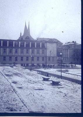 Snow-covered urban plaza with grand neoclassical building featuring twin spires and symmetrical façade. Pedestrians in winter...