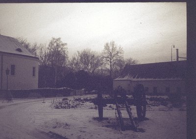 Vintage sepia-toned street scene with light snow covering cobblestone path. Two-story brick buildings flank a narrow road, on...
