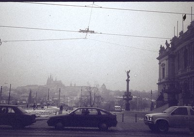 Vintage black-and-white street scene in a European city, likely Prague, showcasing historic architecture with a prominent col...