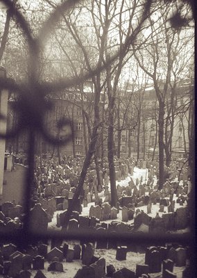 Winter cemetery view through barred window, likely Eastern European. Snow-covered gravestones and leafless trees dominate the...