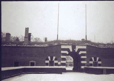 Historic brick fortification entrance with "V" symbol, likely WWII-era German military architecture. Snow-covered ground and ...