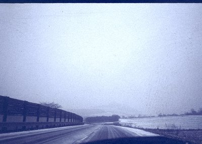 Vintage black-and-white photo of a rural road flanked by snow-covered fields and leafless trees. Industrial brick buildings l...