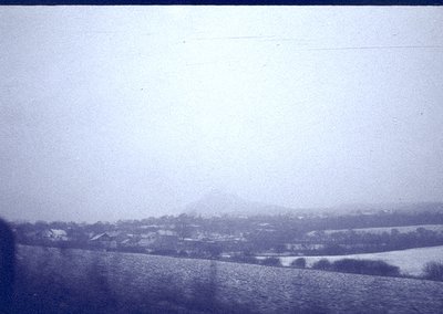 Vintage black-and-white landscape featuring snow-covered fields and distant village. Low-lying hills and misty horizon sugges...